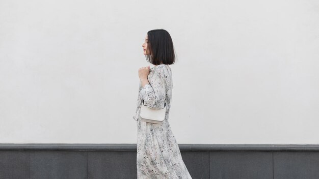 Mooi elegant modemeisje met kapsel in een modieuze bloemenjurk met een stijlvolle tas loopt in de buurt van een witte muur in het zijaanzicht van de stad