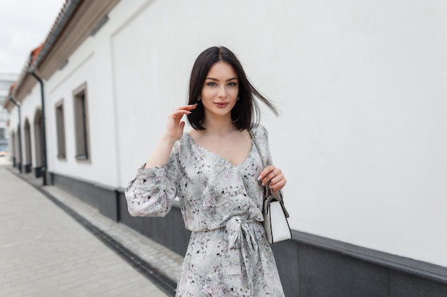 Mooi elegant modieus vrouwenmodel met een bob-kapsel in een stijlvolle zomerjurk met bloemen en een tas die door de stad loopt