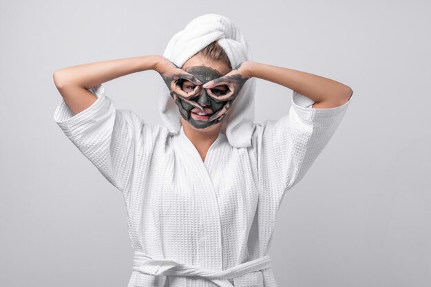 Mooi emotioneel model poseren in een witte badjas met een handdoek op haar hoofd, een hanger om haar nek, met een kleimasker en kleien handen. clownerie naar de camera. een masker maken van vingers.