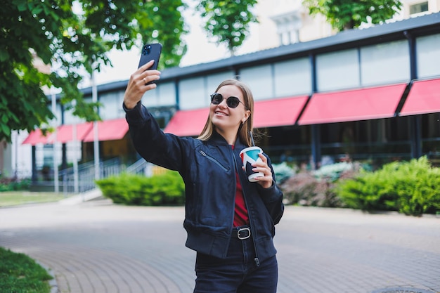 Mooi en jong meisje in zonnebril schrijft een bericht op een mobiele telefoon en buitenshuis koffie drinken