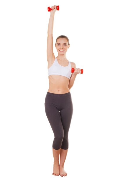 Mooi en sportief. Volledige lengte van mooie jonge vrouw in sportkleding die traint met halters en glimlacht terwijl ze geïsoleerd op wit staat