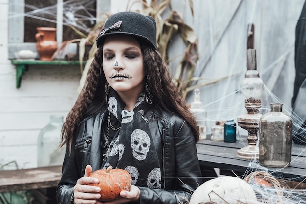 Mooi eng meisje vieren halloween Angstaanjagende zwart witte halfface makeupwitch kostuum stijlvolle afbeelding Horror plezier op kinderfeestje in schuur op straat hoed jas pompoen