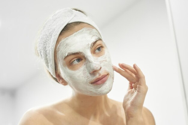 Mooi europees meisje kijkt in de spiegel en brengt een wit masker aan op haar gezicht zomerhuishuidverzorging