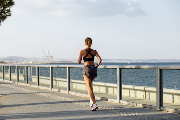 Mooi fitnessmeisje in korte broek en sporttop die bij zonnig weer aan de waterkant aan zee loopt