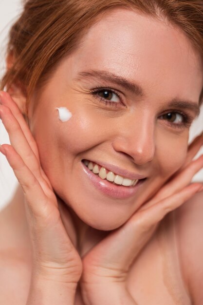 Foto mooi gelukkig jong fris roodharig meisje met een schattige glimlach met crème op haar huidgezicht poses in de studio close-up schoonheid en huidverzorging