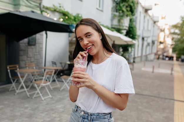 Mooi gelukkig meisje met donker haar met een wit t-shirt drinkt smoothie op straat met groene bomen in het zonlicht op een warme lentedag