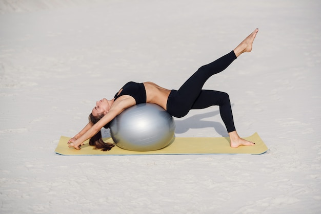 Mooi geschiktheidsmeisje dat uitrekkende oefeningen met geschikte bal op het strand doet. Sport en een gezonde levensstijl concept. Jonge vrouw die pilatesoefeningen doet.