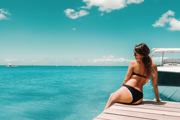 Mooi gevormde brunette in zwembroek en met zonnebril zittend op de pier en genieten van zonnig weer en de oceaan. Teruggedraaid.