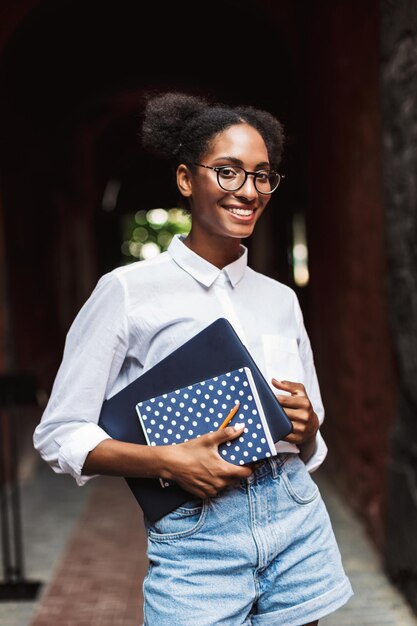 Mooi glimlachend Afrikaans meisje in brillen en shirt met laptop en notitieblok in handen terwijl ze graag buiten in de camera kijkt