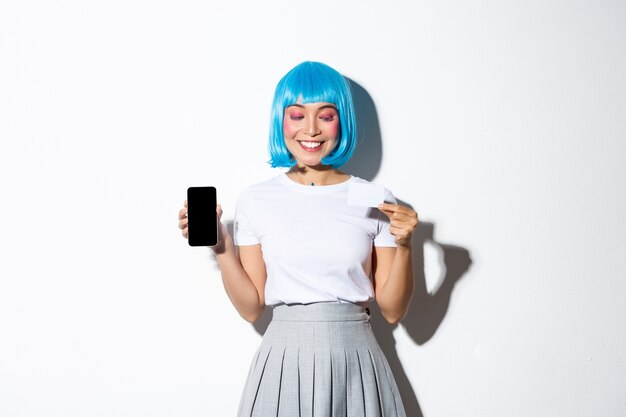 Mooi glimlachend Aziatisch meisje in blauwe pruik die tevreden creditcard bekijkt, het smartphonescherm toont, status.