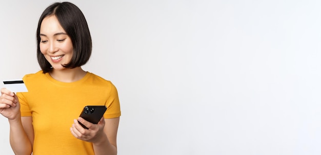 Mooi glimlachend Aziatisch meisje met creditcard en mobiele telefoon die online betaalt op een smartphone die in een gele t-shirt staat op een witte achtergrond