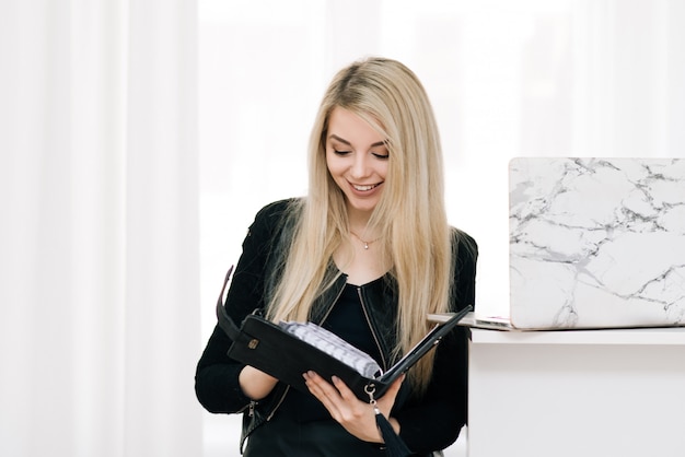 Mooi glimlachend blondemeisje met een notitieboekje en een pen in haar handen, naast laptop op een licht venster, beambte