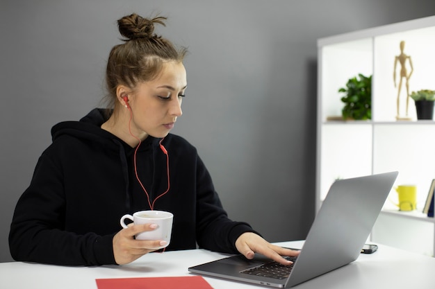 Mooi hipstermeisje die op laptop bloggen die thuis van kop van koffie genieten