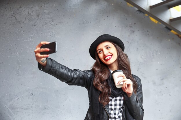 Mooi hipstermeisje met rode lippen en in hoed die selfie in de straat nemen. Kussen verzenden en vredesteken houden. Instagram