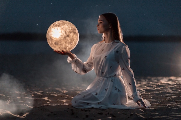 Mooi jong aantrekkelijk meisje op een nachtstrand met zand en sterren houdt de maan in haar handen