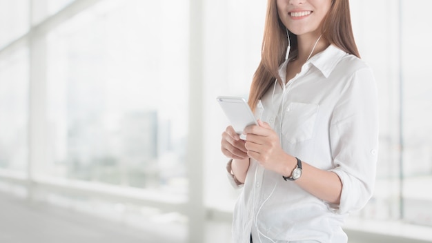 Mooi jong Aziatisch meisje die slimme telefoon met oortelefoon in bureau met behulp van