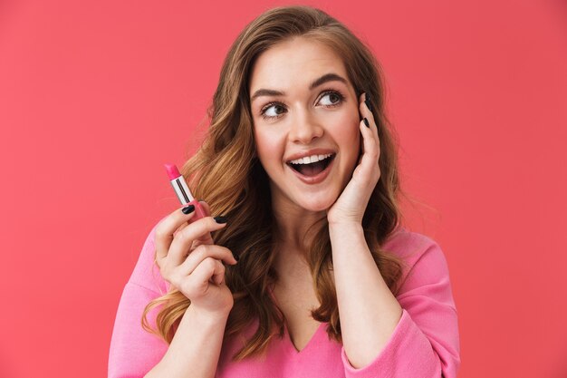 Mooi jong casual blond meisje dat zich geïsoleerd over roze muur bevindt en rode lippenstift vasthoudt