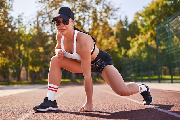 Mooi jong fitness meisje warmt op in het stadion voor de training Aantrekkelijke slanke brunette in een tracksuit zwarte pet en zonnebril Actieve levensstijl