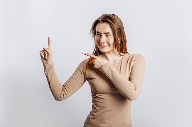 Mooi jong meisje glimlachend en wijzend met haar vingers naar de zijkant op een witte geïsoleerde achtergrond. Een vrouw wijst naar een idee, een plek voor reclame. Positieve brunette in een beige trui.