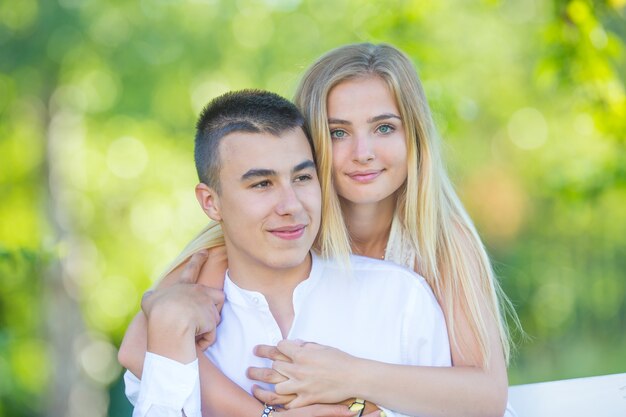 Mooi jong meisje haar donkerharige vriendje van achteren knuffelen terwijl hand in hand in de natuur. Meisje op zoek naar de camera terwijl de jongen op zoek is naar de kant.