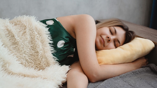 Mooi jong meisje in een groene nachtjapon slaapt op een geel kussen op een bank onder een witte deken