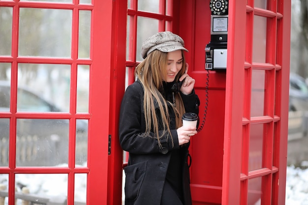 Mooi jong meisje in een telefooncel. Het meisje praat aan de telefoon vanuit de telefooncel. Engelse telefooncel op straat en een vrouw aan de telefoon.