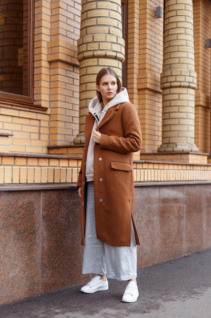 Mooi jong meisje in modieuze biege hoodie, elegante bruine kameeljas en broek op bakstenen muurachtergrond Lopend op straat