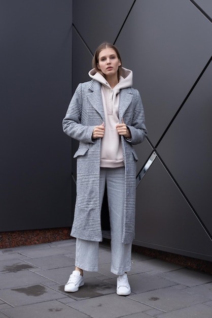 Mooi jong meisje in modieuze biege hoodie elegante grijze jas en broek op donkere zilveren achtergrond Lopend op straat