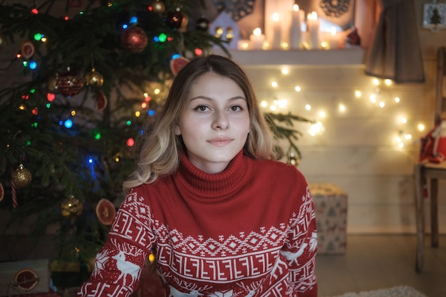 Mooi jong meisje in rode trui op kerstboom. Feestelijk interieur, kaarsen, lichten, slingers. Portret van een meisje op kerstnacht