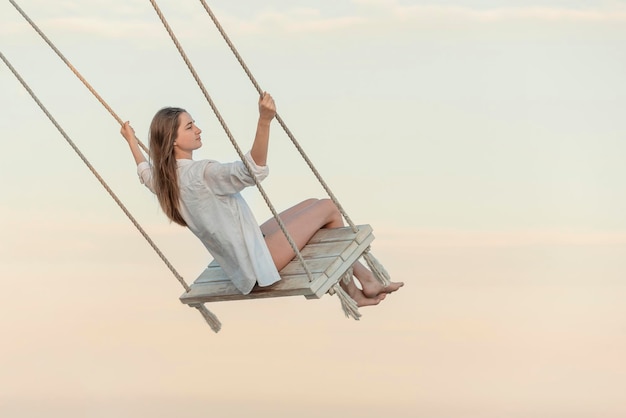 Mooi jong meisje in witte kleren duwt in swing op de lucht achtergrond Mooi zacht zonsondergang licht Mooi meisje zweeft in de lucht