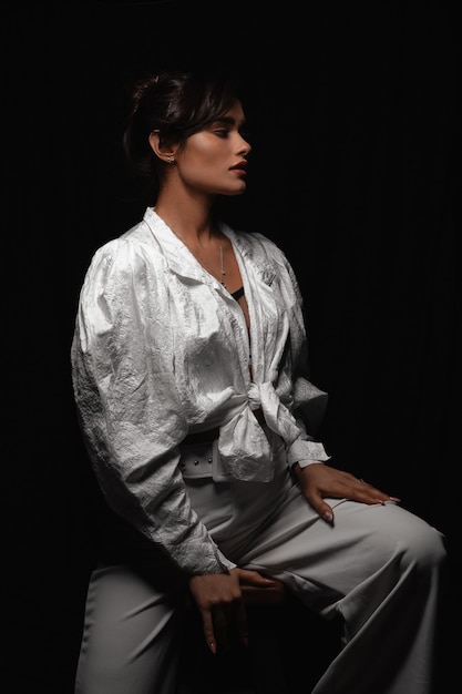 Mooi jong meisje in witte kleren poseren in een donkere fotostudio. Laagdrempelige foto. Gevoelige bruinharige vrouw met lang haar