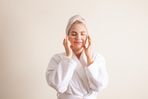 Mooi jong meisje maken zelf gezichtsmassage crème toe te passen met ogen dicht vrouw in witte badjas en handdoek op hoofd dromend poseren op witte achtergrond kopie ruimte