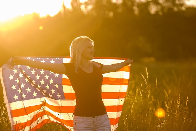 Mooi jong meisje met een Amerikaanse vlag in de wind in een veld met rogge. Zomerlandschap tegen de blauwe lucht. Horizontale oriëntatie.