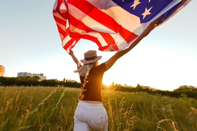 Mooi jong meisje met een Amerikaanse vlag in de wind in een veld met rogge. Zomerlandschap tegen de blauwe lucht. Horizontale oriëntatie.