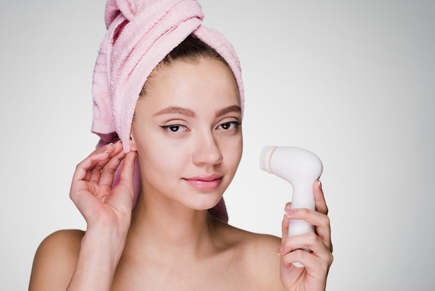 Mooi jong meisje met een roze handdoek op haar hoofd houdt een elektrische borstel vast voor diepe reiniging van het gezicht