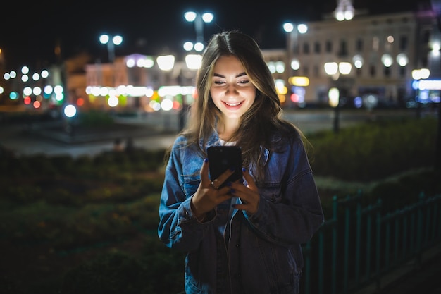 Mooi jong meisje texting op mobiele telefoon buiten over wazig nacht straatmuur, selectieve aandacht