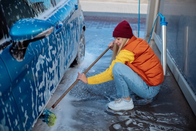 Mooi jong meisje wast de auto bij de zelfbedieningswasstraat. vuil op de auto wissen met een borstel