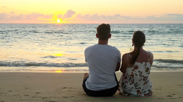 Mooi jong stel zit op het lege oceaanstrand en kijkt naar de zonsondergang in het tropische resort in de avondachterkant