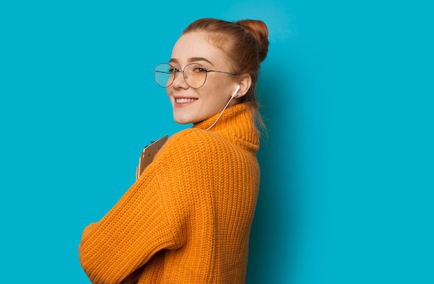 Mooi Kaukasisch meisje met rood haar en sproeten houdt haar laptop vast en gebruikt een koptelefoon op een blauwe muur met vrije ruimte