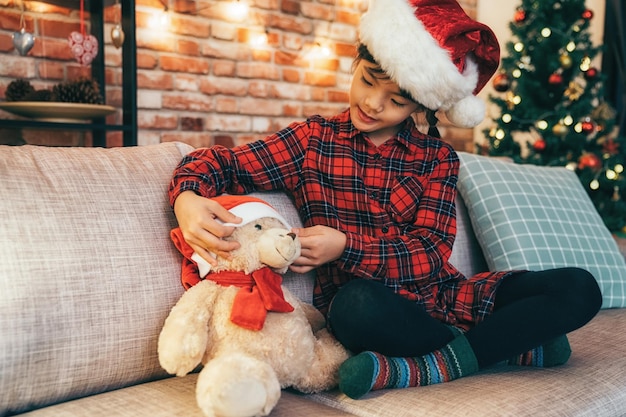 mooi kerstkind in rode hoed zittend op de bank en haar nieuwe speeltje spelend. meisje met haar favoriete teddybeer in de woonkamer. kerstboom en rode bakstenen muur op de achtergrond.