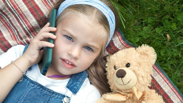 Mooi kindmeisje dat op een deken op groen gazon in de zomer met haar teddybeerstuk speelgoed spreekt op mobiele telefoon.