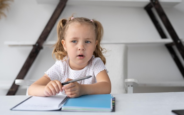 Mooi klein basisschoolmeisje dat haar huiswerk thuis doet terwijl ze aan haar bureau zit met een notitieboekje en pen om te tekenen en handschrift de leerlingschool studeert op afstand