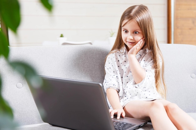 Mooi klein brunette meisje zittend op de bank met een laptopcomputer met een geschokt gezichtsuitdrukkingsconcept voor volwassen inhoud