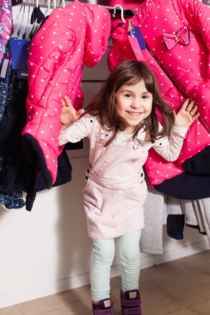 Foto mooi klein meisje dat door kleerhangers met kleren naar buiten gluurt