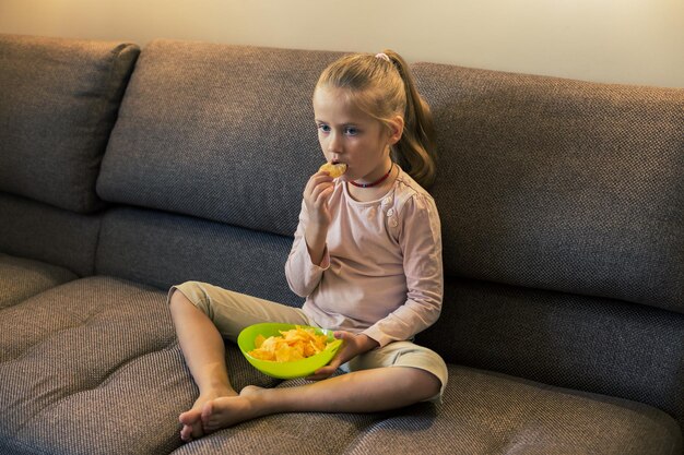 Mooi klein meisje dat ongezond voedsel eet terwijl ze tv kijkt op de bank