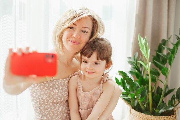 Mooi klein meisje en haar jonge moeder zitten samen en knuffelen, nemen selfies op een rode telefoon