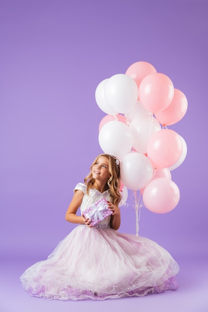 Mooi klein meisje gekleed in prinsessenjurk zitten geïsoleerd over violette muur, met een bos ballonnen en een geschenkdoos