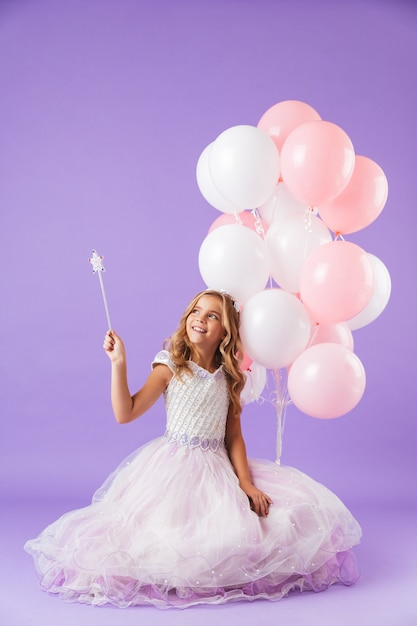 Mooi klein meisje gekleed in prinsessenjurk zitten geïsoleerd over violette muur, met een bos ballonnen en een magische toverstaf