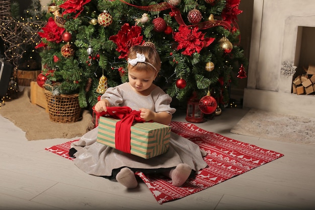mooi klein meisje in een feestelijke jurk zit bij de kerstboom naast een geschenkdoos