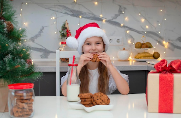 Mooi klein meisje in een kerstmuts drinkt melk en eet koekjes op kerstavond thuis. Het concept van vakantie, Kerstmis en jeugd. Selectieve focus